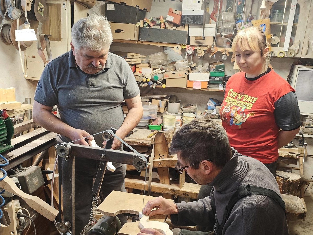Dwóch mężczyzn i kobieta w pracowni stolarskiej, jeden z meżczyzn pracuje przy maszynie, pozostałe osoby obserwują.