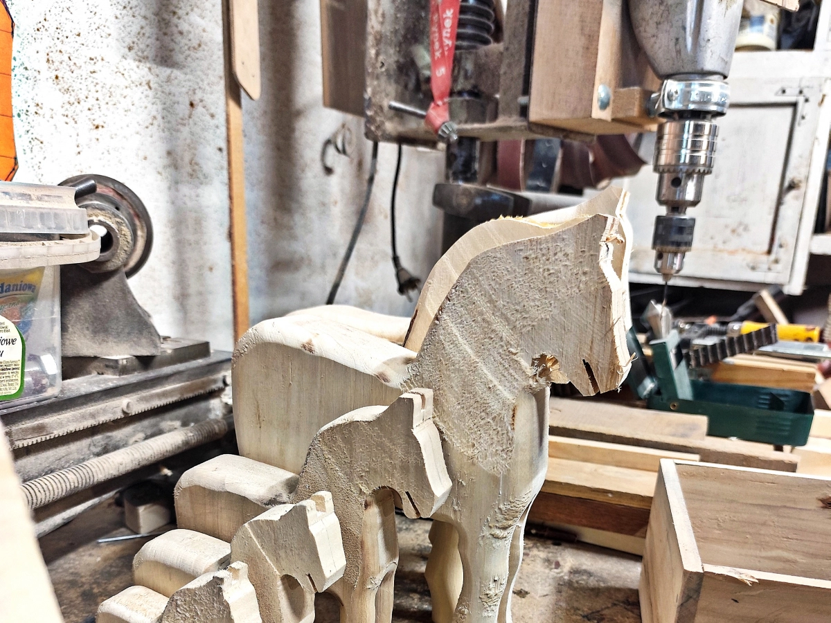 Konie z niewygładzonego drewna różnej wielkości, ustawione w rzędzie na stole w pracowni stolarskiej. W tle maszyny i urządzenia.