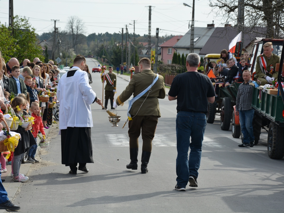Święcenie pokarmów na ulicy: duchowny i uczestnik w mundurze przechodzą obok mieszkańców z koszykami, w tle udekorowane ciągniki.