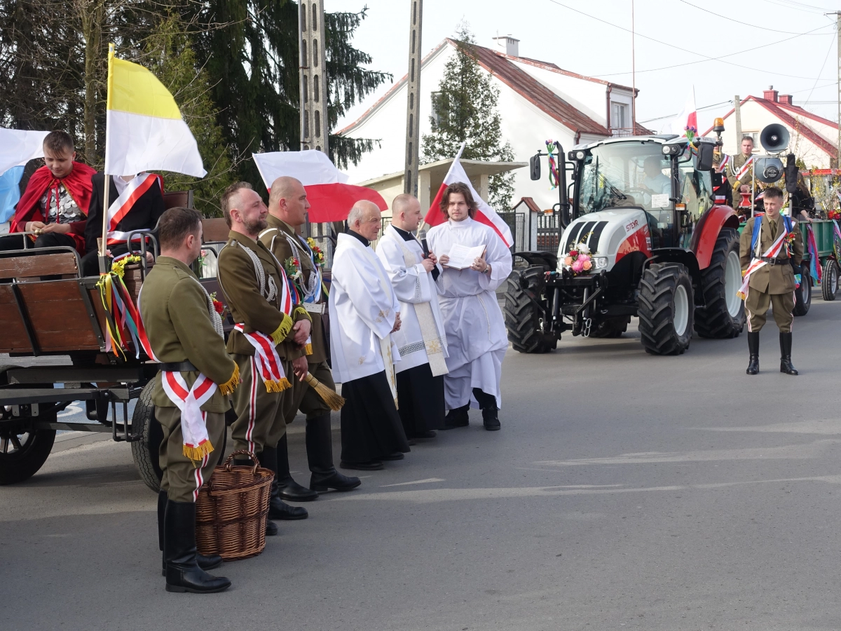 Procesja religijna na ulicy: duchowni i uczestnicy w galowych strojach obok udekorowanych flagami wozów i ciągników.