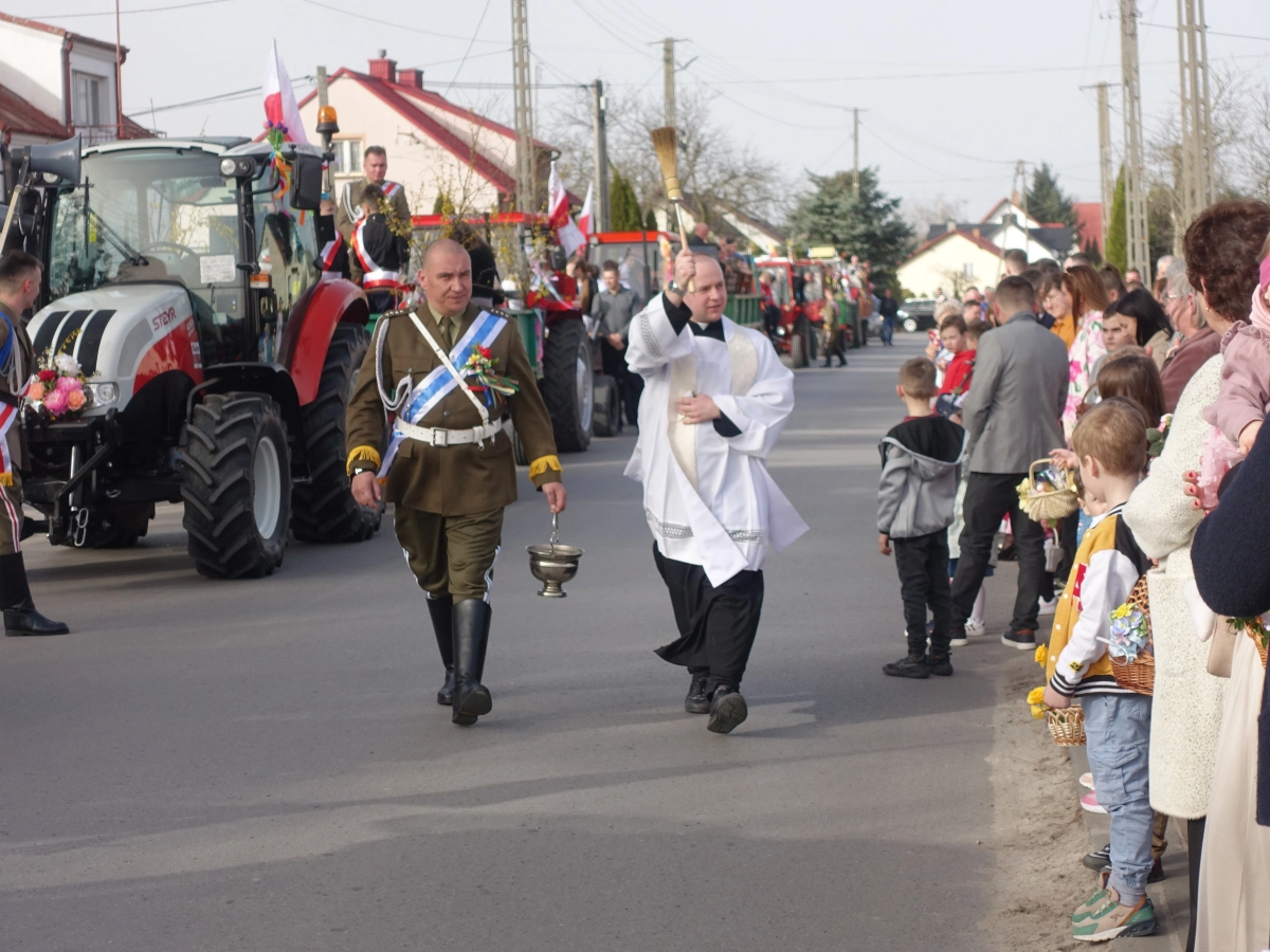 Procesja wielkanocna na ulicy: duchowny kropi wiernych wodą święconą, obok mężczyzna w galowym mundurze; w tle udekorowane ciągniki i mieszkańcy z koszykami.