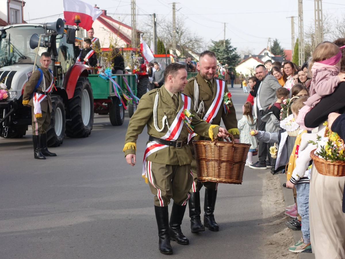 Uczestnicy procesji w galowychch mundurach zbierają dary do kosza od mieszkańców stojących przy ulicy, w tle udekorowane ciągniki.