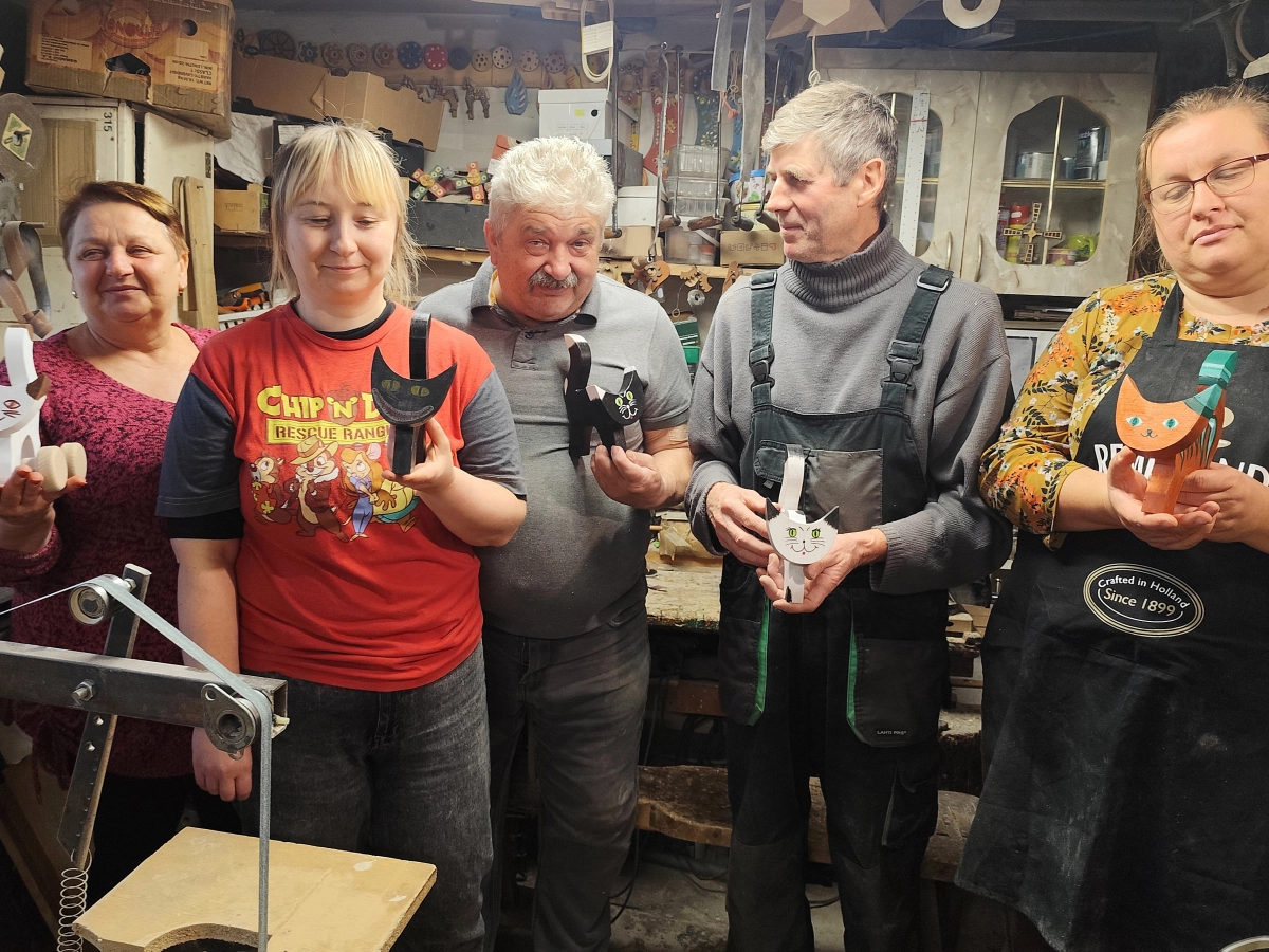 Mężczyzni i kobiety stojąc w grupie prezentują zabawki drewniane - koty, wykonane ręcznie.