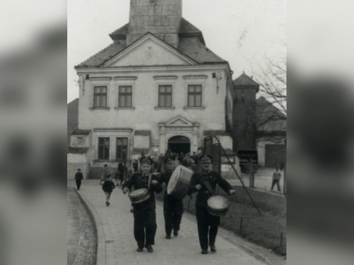 Czarno-białe zdjęcie archiwalne: trzech mężczyzn gra na bębnach, idąc drogą przed budynkiem, z którego wychodzi grupa ludzi.