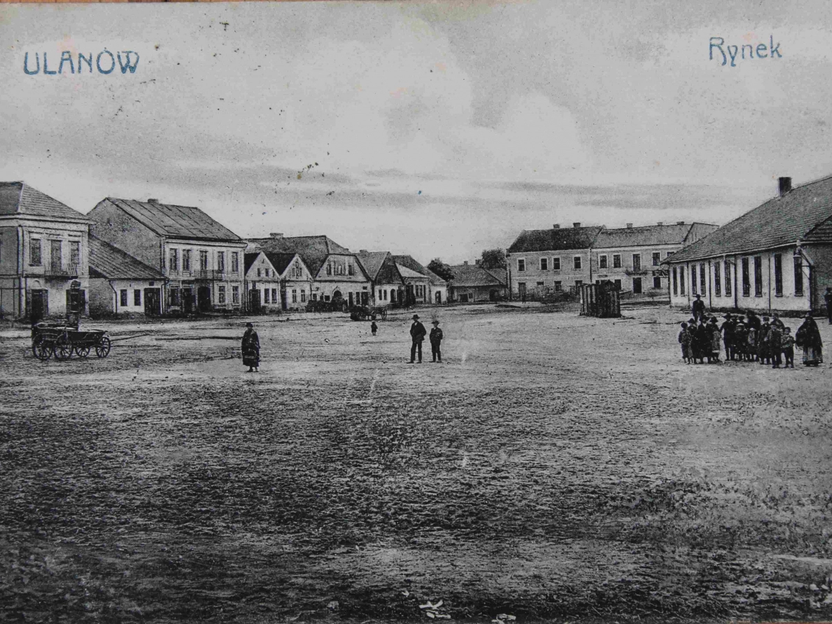 Archiwalna fotografia rynku w Ulanowie. Na rozległym placu stoją mieszkańcy i wóz konny, a wokół rynku znajdują się niskie kamienice i drewniane domy.