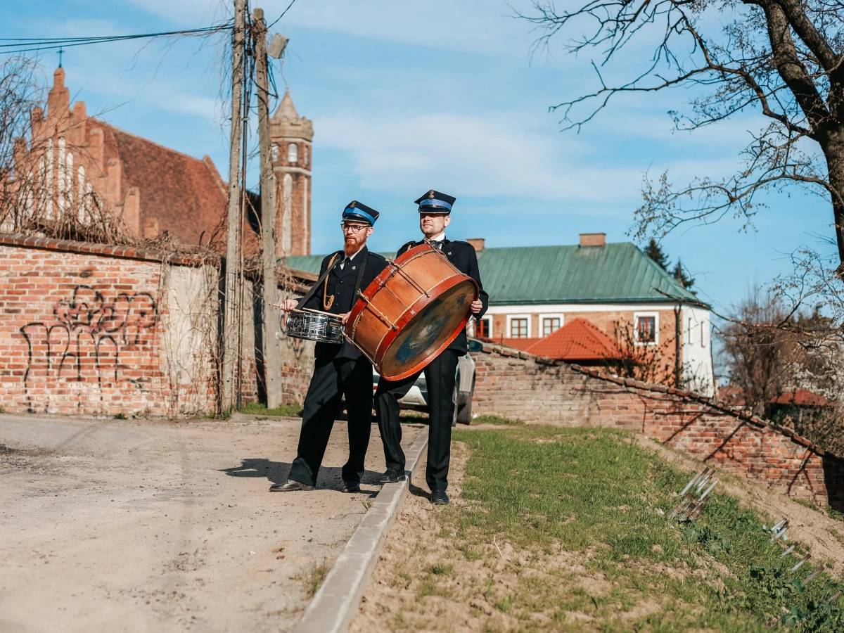 Dwóch mężczyzn w mundurach gra na dużym bębnie i werblu na ulicy, w tle ceglane mury i zabudowania.