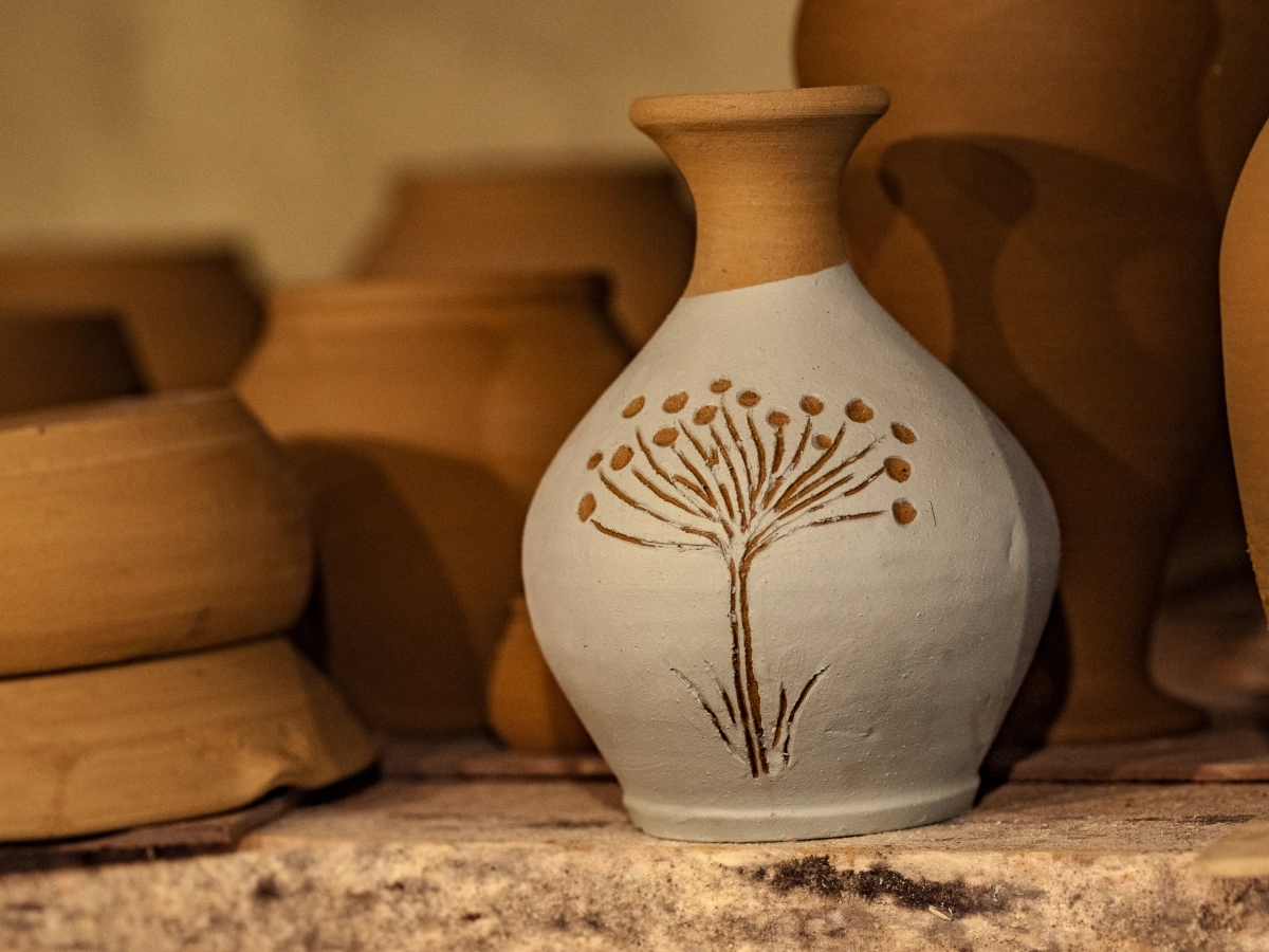 Mały gliniany wazon z roślinnym zdobieniem stoi wśród innych naczyń ceramicznych.