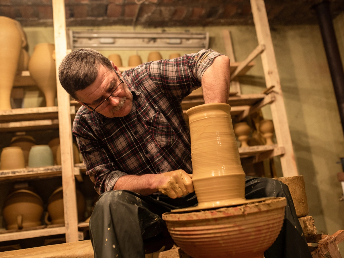 Mężczyzna formuje wysokie naczynie z gliny na kole garncarskim, w tle półki z ceramicznymi wyrobami.