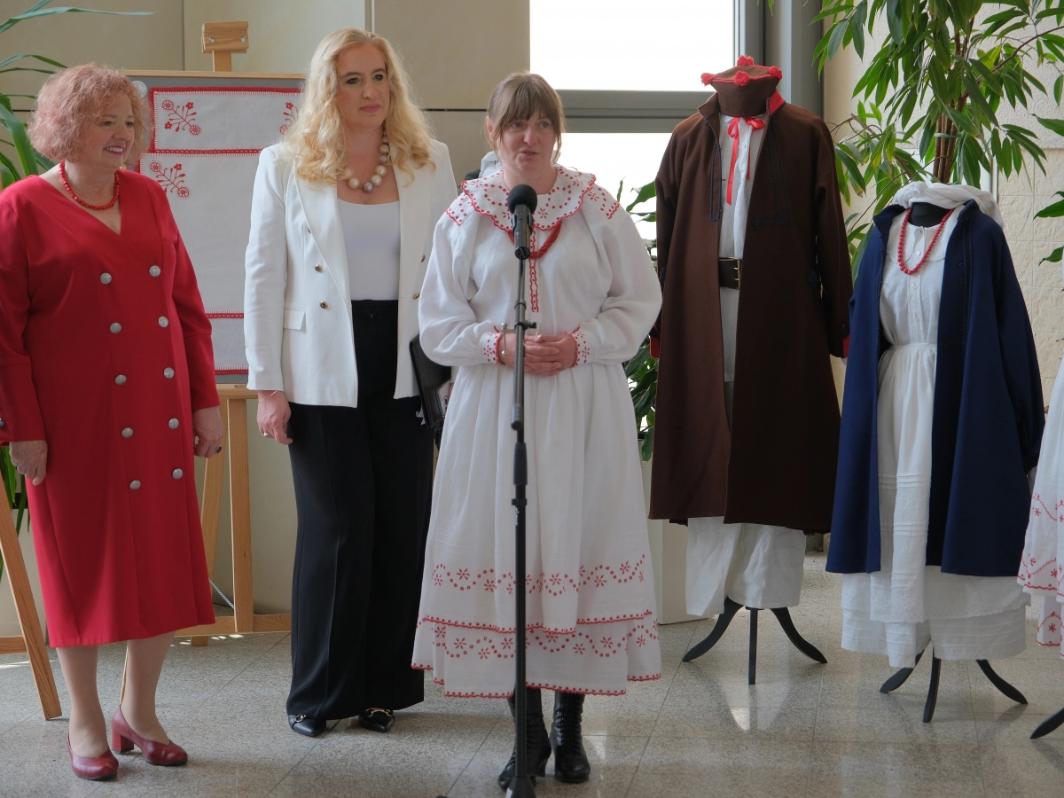 Kobieta w tradycyjnym stroju ludowym przemawia do mikrofonu, stojąc obok dwóch kobiet i manekinów prezentujących regionalne stroje oraz hafty.