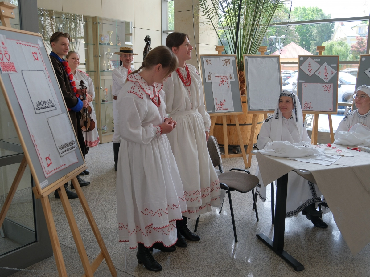 Osoby w tradycyjnych strojach ludowych prezentują regionalny haft, a dwie kobiety siedzą przy stole i wykonują ręczne zdobienia na białym płótnie.