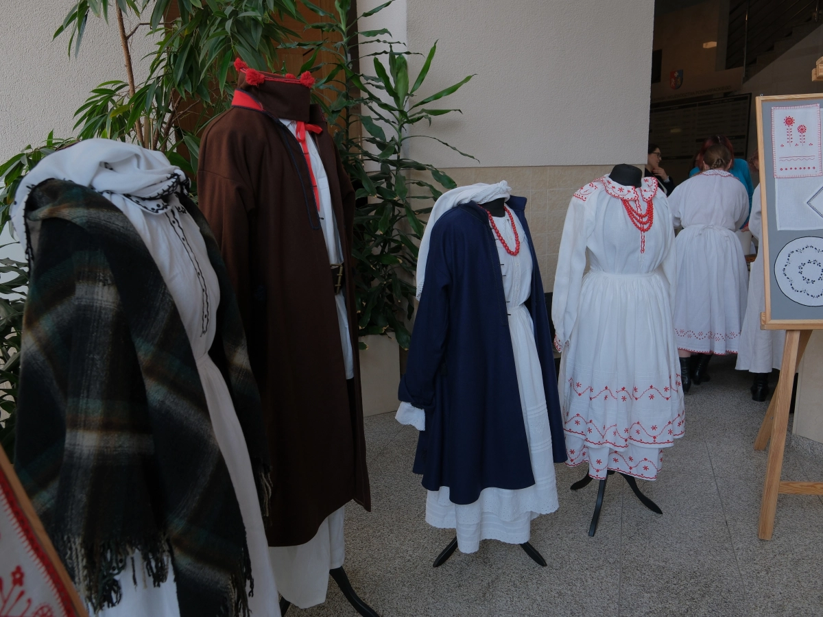 Manekiny prezentują elementy tradycyjnych strojów ludowych z białymi haftowanymi sukniami, granatowym okryciem i brązowym płaszczem. Obok znajduje się plansza z przykładami regionalnych haftów.