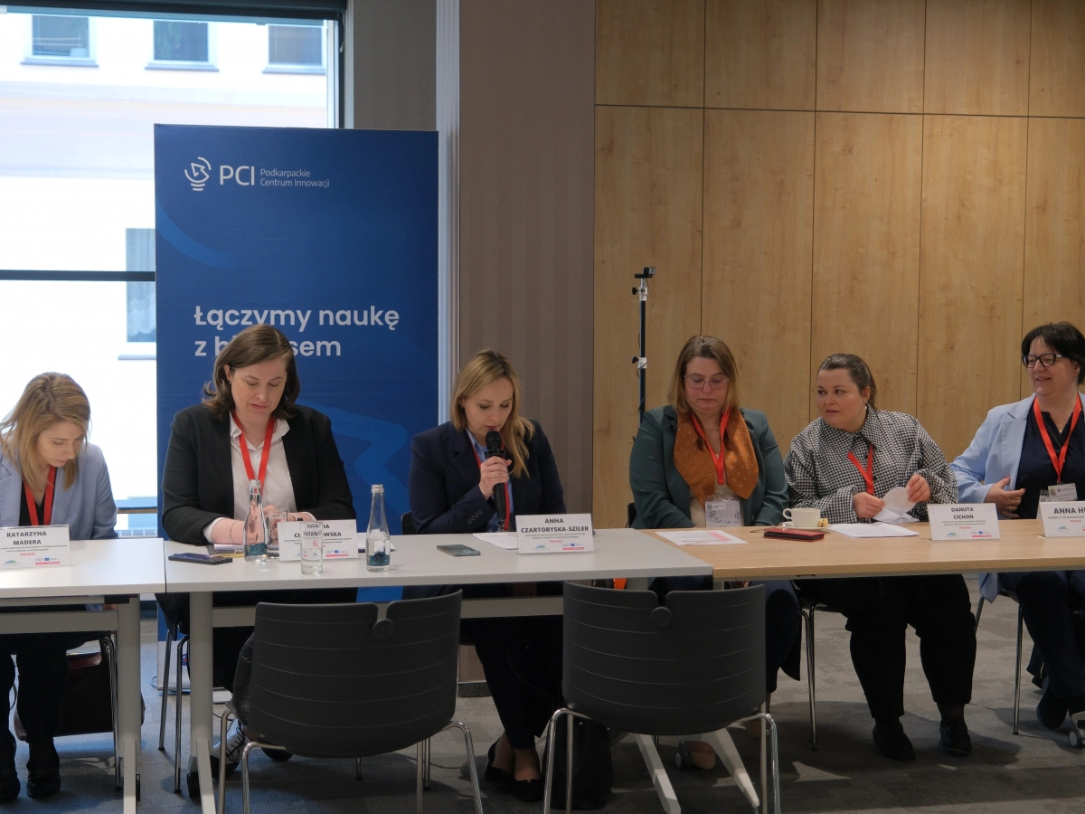 Panelistki siedzą przy stole podczas międzynarodowych warsztatów w Podkarpackim Centrum Innowacji. Jedna z ich przemawia do mikrofonu.