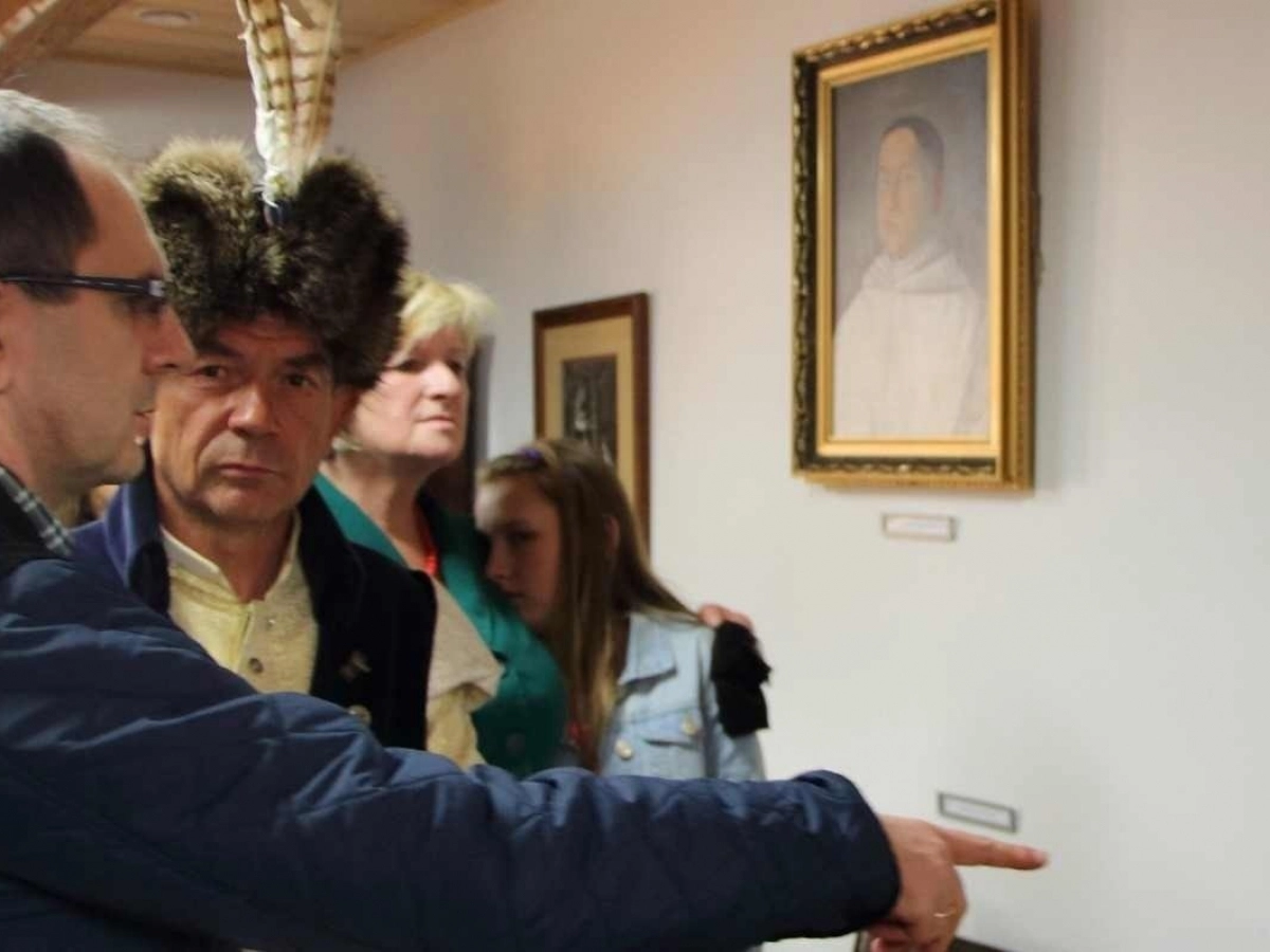 Zwiedzający oglądają ekspozycję w Muzeum Flisactwa Polskiego w Ulanowie. Na ścianie wiszą obrazy, a mężczyzna w tradycyjnej czapce flisackiej rozmawia z uczestnikami.
