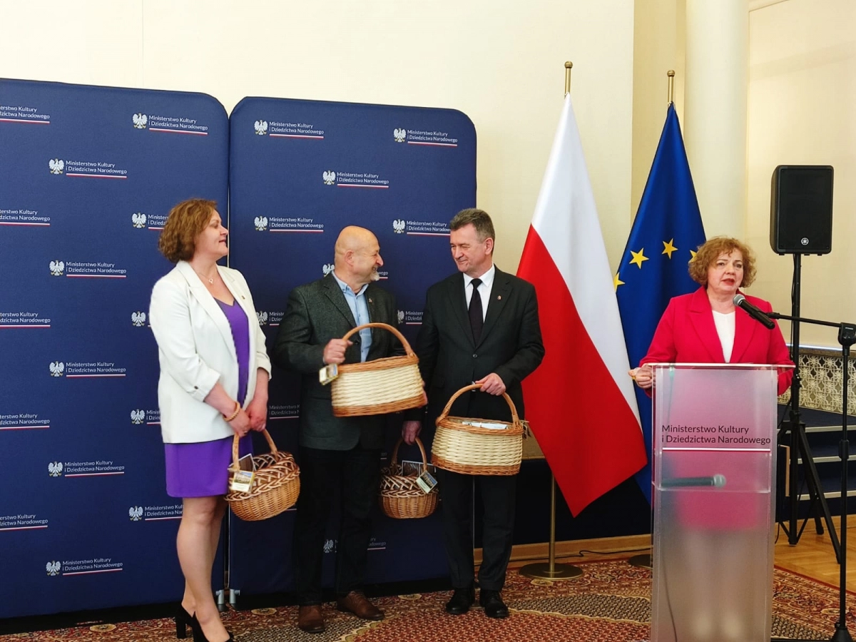 Osoby stojące przed ścianką z logotypami trzymają wiklinowe kosze, kobieta przy mównicy przemawia.