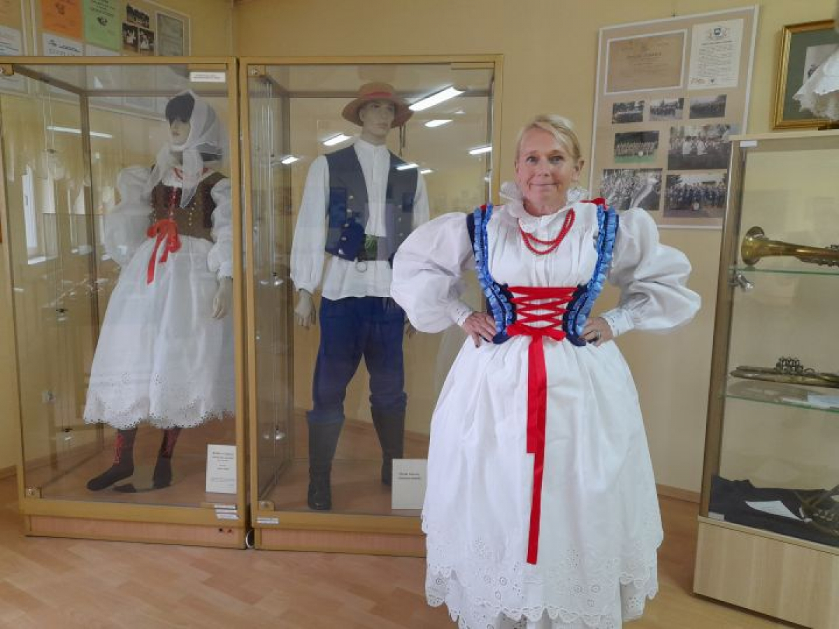 Kobieta prezentuje tradycyjny strój haczowski w muzealnej sali, na tle gablot z historycznymi strojami ludowymi.