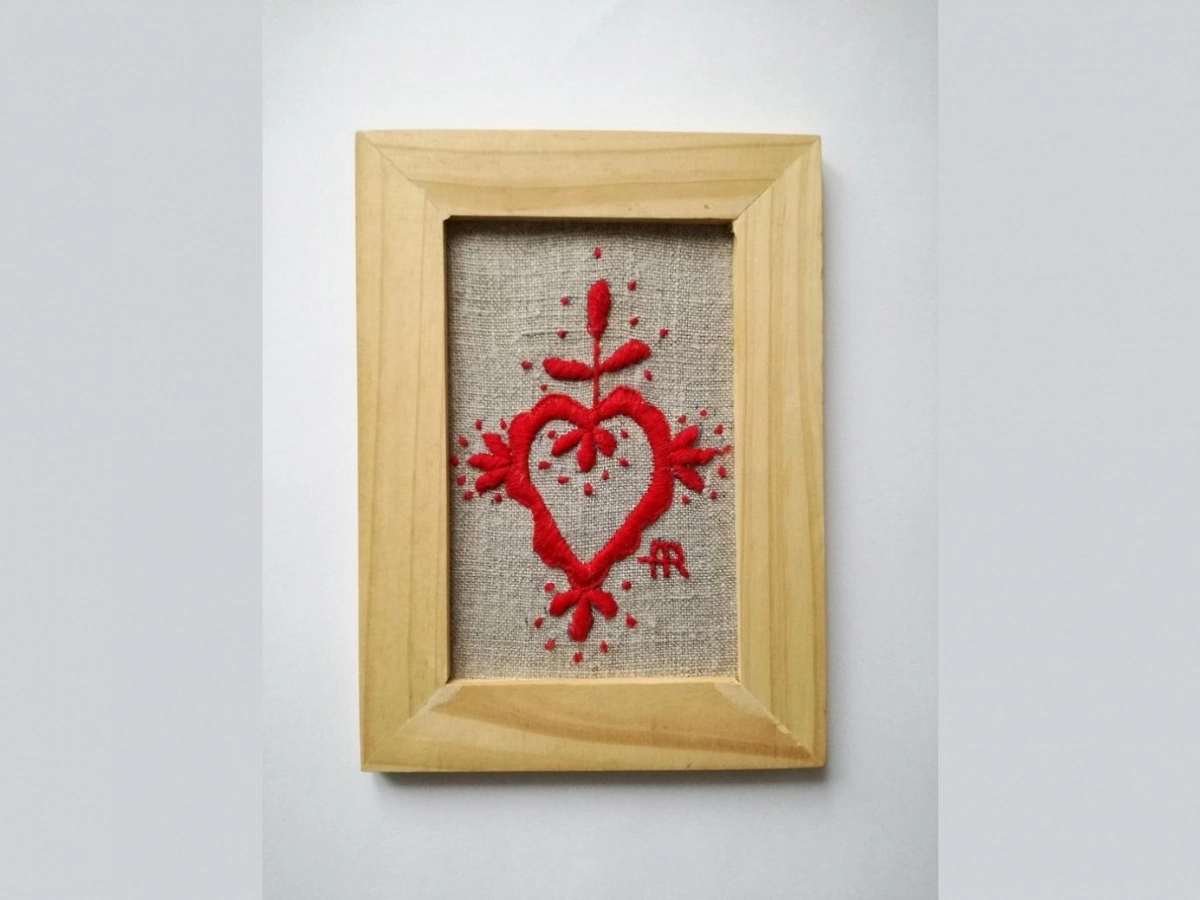 Drewniana ramka z płótnem w środku. Na nim czerwoną nicią wyszyty haft w kształcie serca z ozdobnikami.
