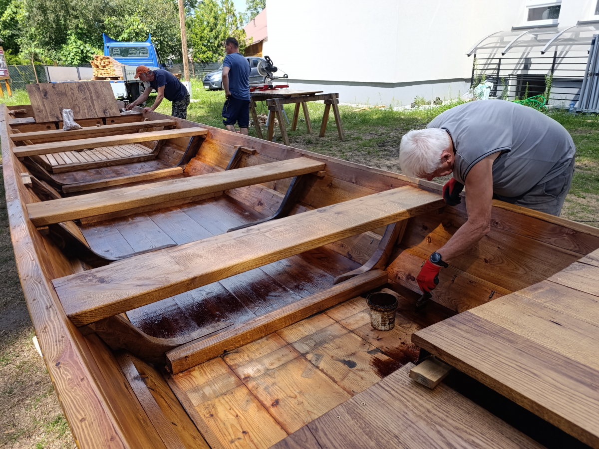 Mężczyźni pracują przy dużej drewnianej łodzi, malując i przygotowując jej wnętrze.