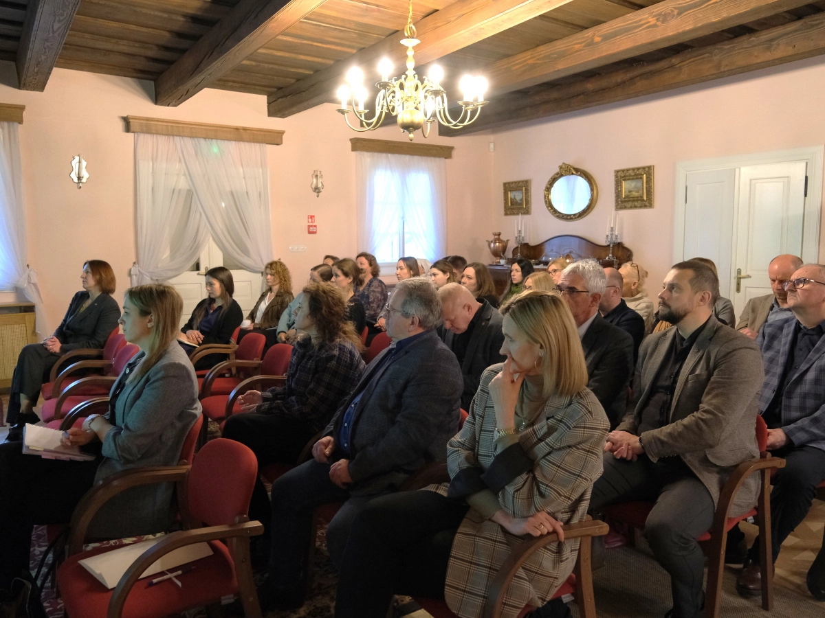 Grupa osób siedzi na krzesłach w sali i uważnie słucha wystąpienia.